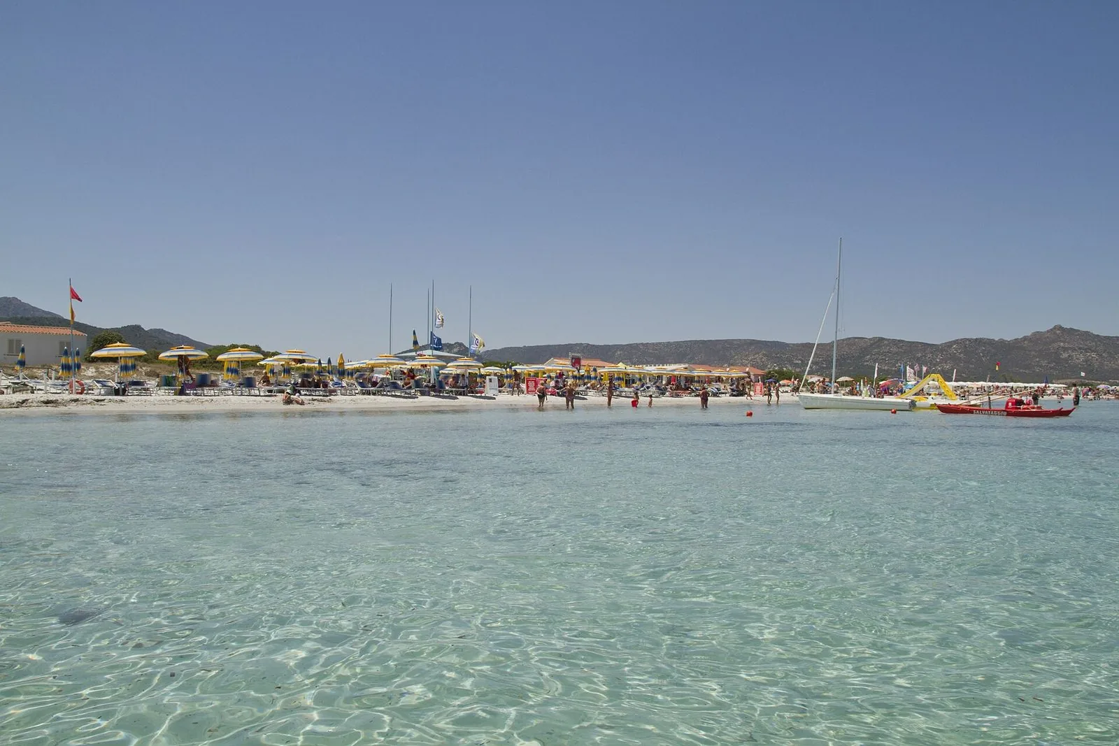 Acqua trasparente e bassa sulla spiaggia La Cinta di San Teodoro in piena estate, con ombrelloni e colline sullo sfondo