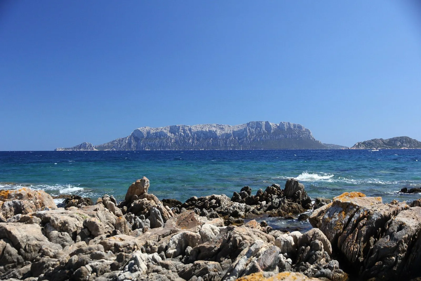 Le pareti calcaree dell'isola di Tavolara viste dalla costa, con scogli in primo piano e mare blu